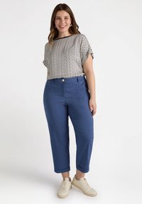 Witte blouse met korte mouwen en een geometrisch patroon, gecombineerd met blauwe tapse broek en beige sneakers. Broek omgeslagen bij de enkels.