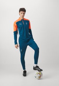 Tuta teal con dettagli arancioni e maniche lunghe, dotata di collo con zip. Il modello si trova in piedi su una palla da calcio, indossando scarpini neri.