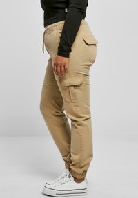 Beige cargo pants with side pockets, elastic cuffs, and adjustable drawstring waist, paired with white sneakers and a black long-sleeve top.