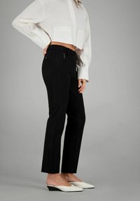 Schwarze maßgeschneiderte Hosen mit schmaler Passform, ausgestattet mit Reißverschlusstaschen vorne und einer glatten Textur. Kombiniert mit einem verkürzten weißen Hemd mit Knopfleiste.