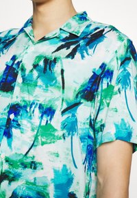 Koszula z krótkim rękawem z lekkiego materiału, z tropikalnym wzorem w niebieskie i zielone palmy na jasnym tle. Pięć guzików.