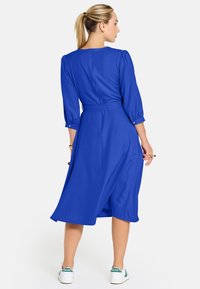 Žena s blond copom, oblečená v modrej midi šatách so zúženým pásom a bielych teniskách, stojí a pozerá sa do strany na jednoduchom pozadí.