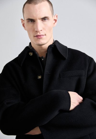 Abrigo de lana negro con diseño de cuello, cierre de botones y un bolsillo lateral. Textura suave con un ajuste holgado.