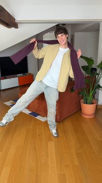 Osoba u bež jakni preko bijele majice, sivim trapericama i srebrnim cipelama, držeći tamnoljubičasti šal, stoji na drvenom podu.