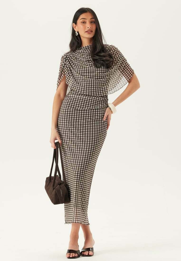 Femme en robe à carreaux noir et blanc avec un voile transparent, tenant un sac à main marron foncé, portant des talons noirs et un bracelet blanc.