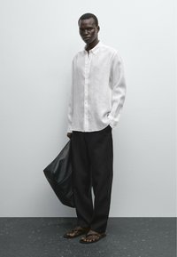 White long-sleeve linen shirt, black loose trousers, and black flip-flops. A large black bag is held in the left hand. Plain background.