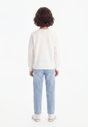 Enfant aux cheveux bruns bouclés, debout de dos, portant un t-shirt blanc à manches longues, un jean bleu clair et des baskets blanches sur fond blanc.