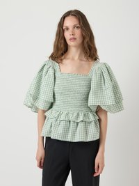 Blusa verde a quadretti con maniche a sbuffo, scollatura quadrata, corpetto arricciato e orlo a volant. Indossata con pantaloni neri. Tessuto leggero.