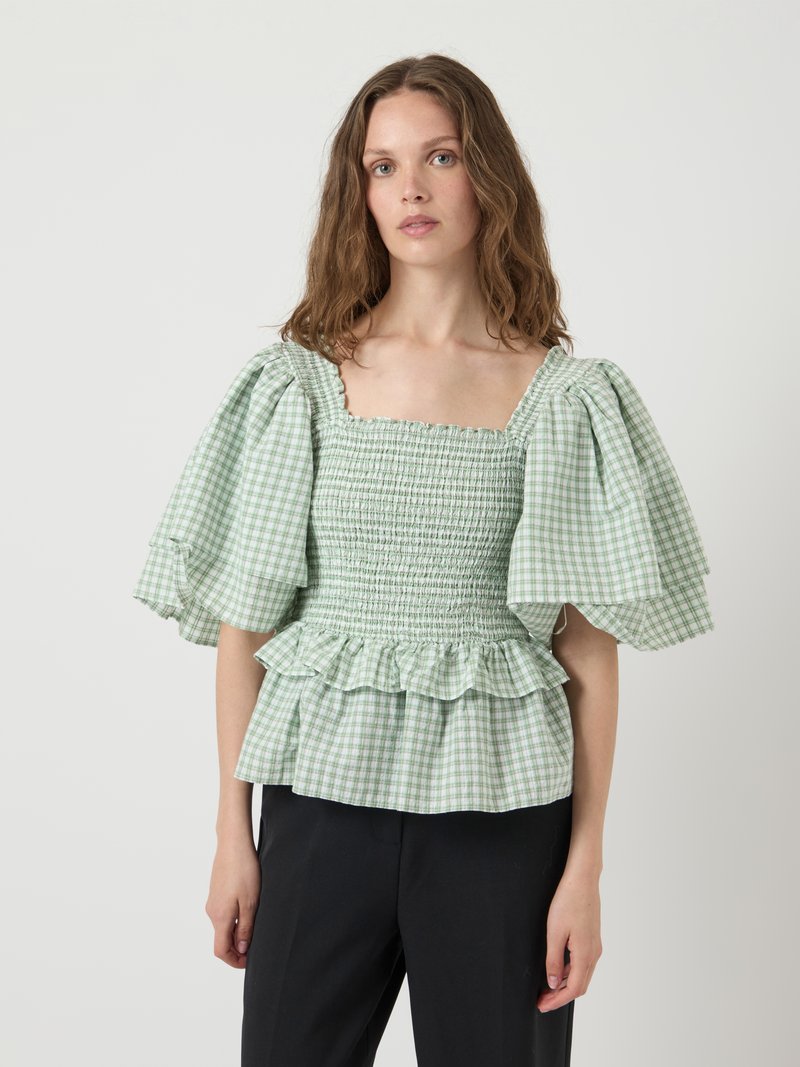 Blusa verde a quadretti con maniche a sbuffo, scollatura quadrata, corpetto arricciato e orlo a volant. Indossata con pantaloni neri. Tessuto leggero.