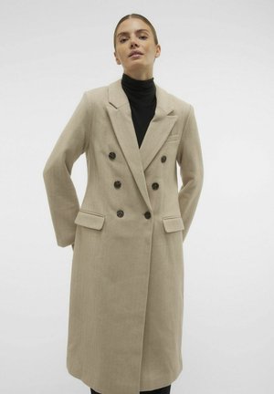 Beige dubbelzijdig jasje met visgraatstructuur, grote lapels, vier knopen en twee voorkruiden. Draagt over een zwarte hoge hals top.
