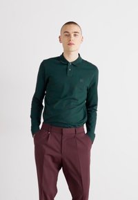 Cămașă polo cu mânecă lungă de culoare verde închis, cu guler, asortată cu pantaloni maronii cu talie înaltă, având un design pliat și buzunare laterale.