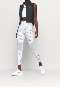 Chaleco negro acolchado sobre un top corto blanco, combinado con leggings ajustados blancos con un patrón de salpicaduras negras y detalles de malla. Zapatillas blancas.