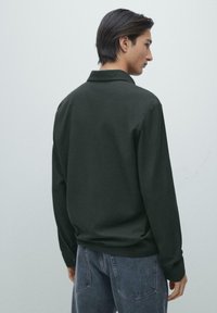 Dunkelgrünes Polohemd mit langen Ärmeln und strukturierter Stoffqualität, ausgestattet mit einem klassischen Kragen und einer Knopfleiste, kombiniert mit hellblauen Jeans.