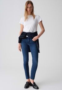 T-shirt blanc à manches courtes associé à un jean skinny bleu foncé, veste en denim nouée à la taille et mocassins noirs. Tenue simple et décontractée.