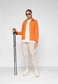 Orange Fleecejacke mit Reißverschluss, beiger T-Shirt, beige Jogginghose und hellen Sneakers. Hält schwarze Trekkingstöcke mit ergonomischen Griffen.