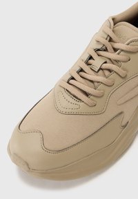 Beige sneaker made of textured fabric with a smooth rubber sole. Features laces, curved lines, and subtle stitching details.