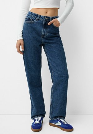 Blaue hochgeschnittene Jeans mit lockerer Passform, subtilen Auswaschungen, fünf Taschen und einem Knopfverschluss. Kombiniert mit blauen Turnschuhen und einem weißen Langarmshirt.