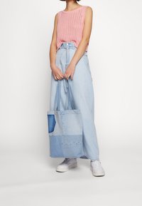 Débardeur en maille rose clair à coupe ample, associé à une longue jupe en denim bleu clair avec des accents en patchwork. Baskets blanches et sac fourre-tout.