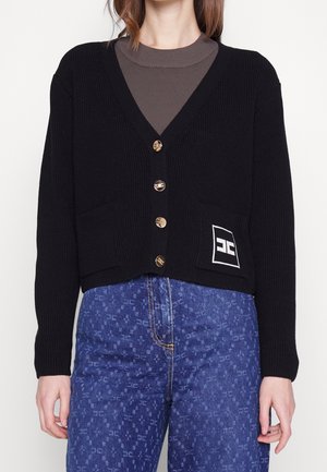 Kobieta ubrana w czarny sweter na guziki z naszywką na kieszeni, na szarej bluzce, do tego niebieskie jeansy w wzory.