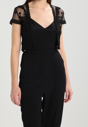 Kvinde iført sort ærmeløs jumpsuit med en gennemsigtig, kortærmet, blomsterbroderet blonder shrug over skuldrene.