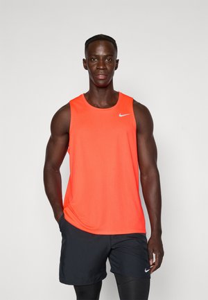 Homme athlétique debout, les mains dans les poches, portant un haut sans manches orange vif et un short noir par-dessus des leggings noirs.