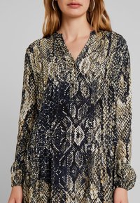 Šaty s dlouhým rukávem s hadím vzorem v tmavě modré, krémové a zlaté barvě. Měkká látka s výstřihem do V a nařasenými rukávy.