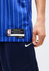 Μπλε φανέλα NBA με λευκές κάθετες ρίγες και ετικέτα μεγέθους 44, φορεμένη με σκούρα μπλε σορτς Nike που φέρουν λευκό λογότυπο swoosh.