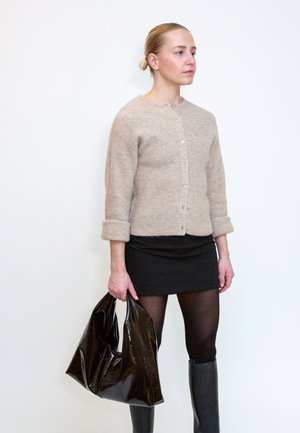 Femme debout portant un cardigan beige, une mini-jupe noire, des collants noirs et des bottes noires montant jusqu’aux genoux, tenant un grand sac noir brillant.