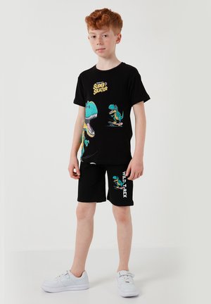 Niño pelirrojo con camiseta negra y pantalones cortos con gráficos coloridos de T-Rex en monopatín y zapatillas blancas, posando sobre un fondo liso.