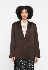 Brūns pielāgots žakete ar platiem atlokiem, divām priekšējām kabatām, pogu aizdari un gludu audumu. Valkāts pār krēmkrāsas topu un melnu svārku ar baltām punktiņām.