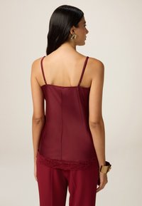 Burgundy camisole, jossa on ohuet olkaimet ja pitsireunus helmassa, valmistettu sileästä kankaasta, kuvattu takaa.