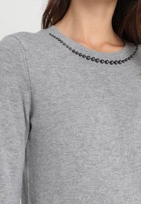 Pull gris en tricot avec un col rond, doté d'une rangée d'ornements foncés autour du col pour plus de détails.