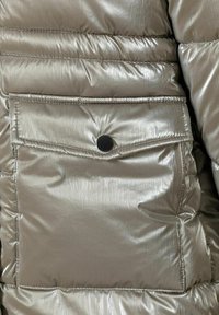 Gros plan sur une doudoune argent métallique présentant une surface texturée, des coutures horizontales et une poche à rabat avec un bouton pression noir.