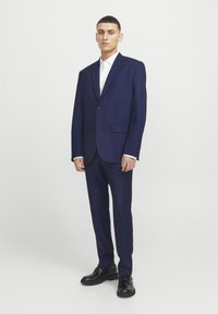 Chaqueta y pantalones de traje azul marino con un solo botón, bolsillos y una textura suave, combinados con una camisa blanca y zapatos negros.