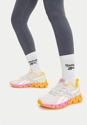 Zapatillas deportivas blancas con suelas de goma rosa y naranja, parte superior de malla y detalles con logotipos. Se llevan con mallas grises y calcetines deportivos blancos.