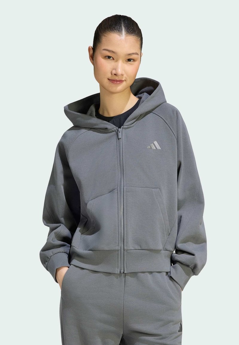 Jeune femme portant un sweat à capuche gris zippé et un pantalon de survêtement assorti avec des logos Adidas, posant les mains dans les poches sur un fond clair.
