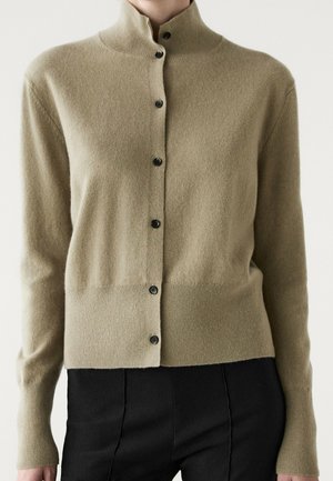Hellbrauner, geknöpfter Cardigan mit hohem Kragen, über schwarzen Hosen getragen, von den Schultern bis zu den Hüften gezeigt.
