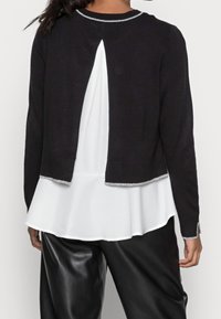 Black cardigan featuring a partially open back, silver trim at neckline and hem, layered over a white blouse with a flowing silhouette.