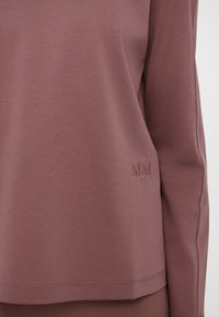 Piedurkņu topi ar garām piedurknēm izgatavots no putekļainā mauve auduma ar gludu tekstūru. Iezīmēts ar smalku izšuvumu "MM" apakšējā labajā stūrī.
