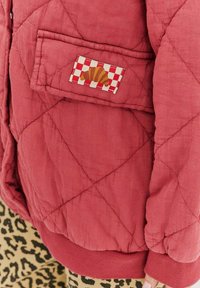Prešívaná červená bunda s vreckom so štvorcovou nálepkou a štýlovým dizajnom, s rebrovaným lemom a hladkou textúrou.