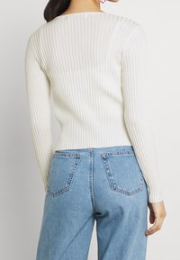 Žebrovaný bílý svetr s dlouhým rukávem a přiléhavým pasem, zkombinovaný s světle modrými džínami s kapsami na zadní straně a volnějším střihem.