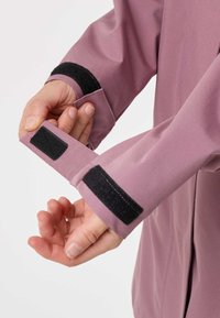 Ruky upravujú manžetu mauve bundy s čiernymi suchými zipsami na bielom pozadí.