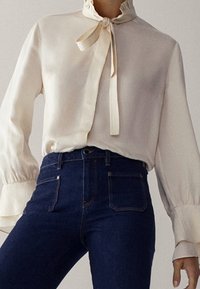 Krämfärgad blus med hög volangkrage och fluga, tillsammans med högmidjade mörk denimjeans med framfickor och en figurnära silhuett.