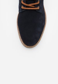 Zapato de ante azul marino con cordones marrones, costuras en contraste y suela de goma marrón. Presenta una puntera redondeada y una superficie texturizada.