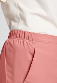 Pantaloni in tessuto rosa, morbido al tatto, con cintura elastica e tasche laterali piatte, mostrati di lato su uno sfondo chiaro.