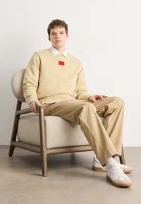 Maglione beige con etichetta rossa, camicia bianca con colletto, pantaloni marrone chiaro, sneakers bianche, seduto su una sedia di legno davanti a uno sfondo semplice.