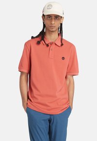 Korallfärgad pikétröja med krage och tre knappar, med en liten svart logotyp på vänster bröst. Kombinerad med blå shorts, händerna i fickorna.