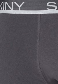 Primo piano di un tessuto grigio scuro con cuciture visibili e una parte di vita elastica etichettata "SKINY" in testo bianco.