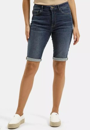 Kobieta nosząca podwinięte, ciemnoniebieskie bermudy z denimu, zestawione z białymi espadrylami na platformie bez sznurowadeł.