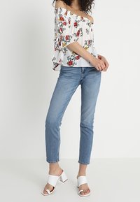 Vit off-shoulder topp med blommönster, flaggande ärmar och snördetaljer. Matchas med ljusblå skinny jeans och vita blockklack-sandaler.
